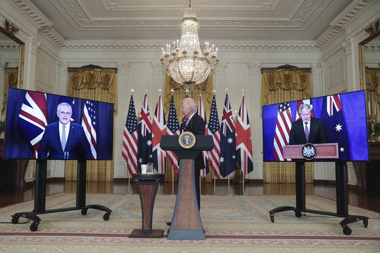 man at lecture between two TV screens displaying men's faces