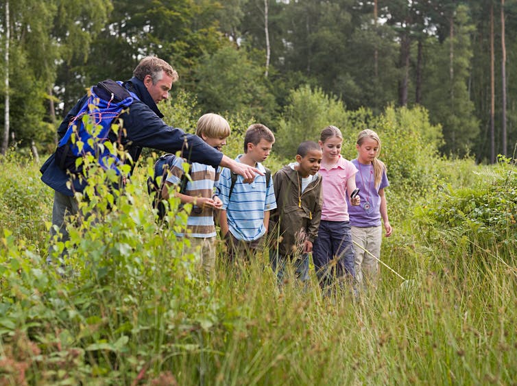A man points into a pond where 5 kids are looking with excited expressions on their faces.