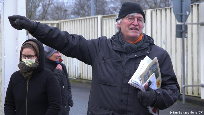 a man with gray hair and holding papers gestures with his hand as people stand behind