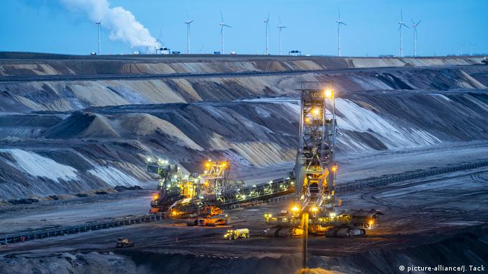 Sprawling landscape of a lignite mine, with machinery in the middle and wind turbines in the background