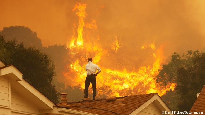 A man stands on the roof of a house looking a raging wildfire. 