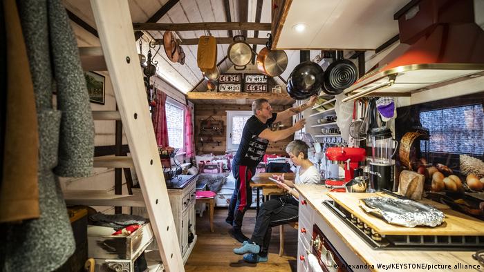 Two people inside of a tiny house where kitchen utensils hang from the wooden ceiling