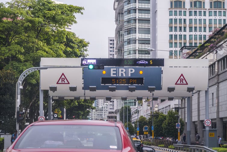A car approaches an overhead billboard displaying time of day and prices for cars and trucks to enter the regulated zone.