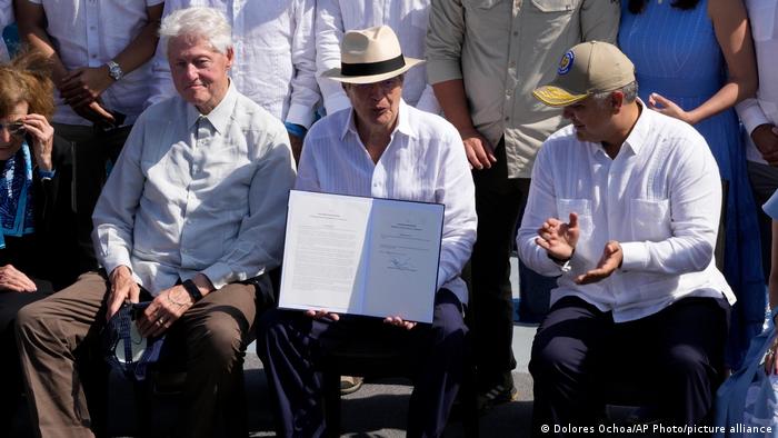 Three men: Bill Clinton with ball cap in hands, Guillermo Lasso in a brimmed hat, Duque
