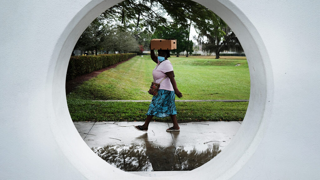 woman wearing mask, carrying cardboard box on her head, seen through a round window
