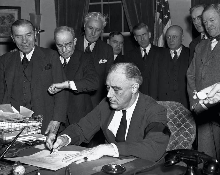 A crowd of men gathers behind President Franklin D. Roosevelt as he signs a paper.