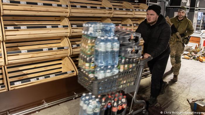 Two men walk down an aisle with empty shelves, pushing a cart stacked with water and soft drink bottles 