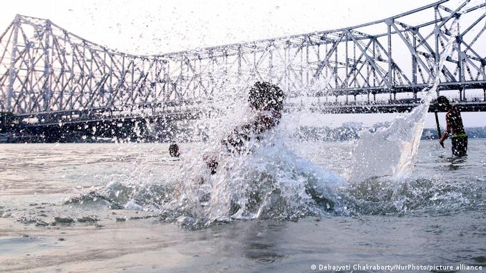 A boy jumps in a river near a large steel frame bridge
