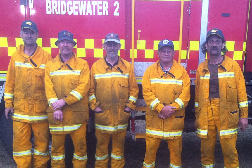 Five uniformed firefighters standing in front of a fire truck.
